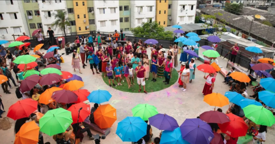 Foto de diversas pessoas reunidas na área externa de um conjunto habitacional. Elas formam uma roda, com diversas pessoas usando roupas coloridas e portando guarda-chuvas nas cores azul, roxo, verde, vermelho, vinho e laranja. No centro da roda, um jovem branco de cabelos escuros, camiseta vermelha e bermuda branca fala e gesticula. Ao lado dele há crianças e jovens, muitos deles negros. Ao fundo, parte da fachada de alguns prédios. 