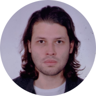 foto em primeiro plano de um homem branco de olhar sério. Ele tem cabelos castanhos, lisos e na altura do ombro e veste uma camiseta branca e blusa preta. O fundo é branco.