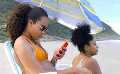 Imagem do filme &quot;Receita de Caranguejo&quot;. Há, na foto, duas mulheres pretas em uma praia. A mulher em primeiro plano, sentada e usando óculos e biquíni amarelo, olha para seu celular de capa laranja. A outra, de biquíni e brinco azul, olha para a paisagem. Ao fundo, pode-se observar uma montanha e parte do mar.