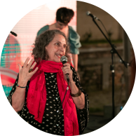 Foto de mulher falando ao microfone e gesticulando. Ela tem cabelos ondulados e castanhos até os ombros, veste uma camisa de mangas largas com detalhes dourados e um cachecol vermelho. No fundo há um palco com microfone e uma pessoa
