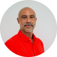 Foto de homem branco esboçando um sorriso. Ele é careca, tem bigode grisalho e olhos castanhos. Usa uma camisa vermelha. O fundo é cinza. 