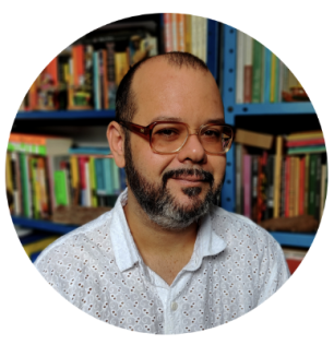 Foto de homem branco de semi-perfil. Ele tem cabelos, barba e bigode bem curtos, pretos e um pouco grisalhos. Usa óculos e camisa branca. No fundo desfocadas, estantes cheias de livros.