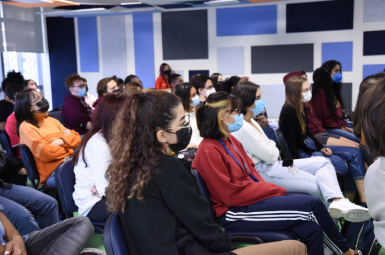 Foto de vários jovens sentados em cadeiras azuis concentrados assistindo à palestra, todos usam máscara. A parede ao fundo tem formas geométricas azuis como decoração.