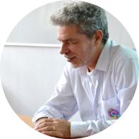 Foto de homem branco de perfil com braços apoiados em uma mesa. Ele tem cabelos curtos, ondulados e grisalhos e barba aparada e branca. Ele veste camisa social branca. Ao fundo, uma parede branca com um quadro branco.