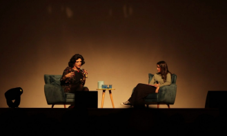 Foto de Rita e Amanda sentadas em cadeiras aveludadas sobre o palco. A iluminação do ambiente é baixa. Rita, que gesticula e segura um microfone, está à esquerda, é branca, tem cabelos escuros médios e veste uma roupa florida. Amanda, que olha para a convidada, tem cabelos escuros longos e lisos, é branca e veste uma blusa cinza, calças pretas e tênis.