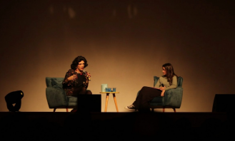 Foto de Rita e Amanda sentadas em cadeiras aveludadas sobre o palco. A iluminação do ambiente é baixa. Rita, que gesticula e segura um microfone, está à esquerda, é branca, tem cabelos escuros médios e veste uma roupa florida. Amanda, que olha para a convidada, tem cabelos escuros longos e lisos, é branca  e veste uma blusa cinza, calças pretas e tênis.