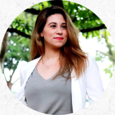 Foto de uma mulher branca de cabelos compridos, lisos e descoloridos nas pontas. Ela usa blusa cinza, blazer branco e colar prateado. Atrás dela há folhas e galhos de uma árvore.