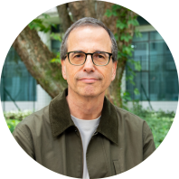Foto de homem branco. Ele tem olhos castanhos, cabelos curtos e grisalhos, usa óculos de aro retangular e camisa branca sob uma jaqueta verde musgo. Ao fundo, folhagens, uma árvore e janelas de um prédio.