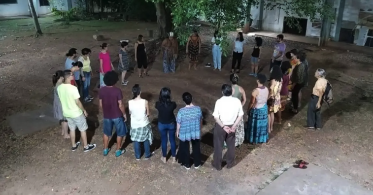 Foto de uma roda de diversas pessoas, homens e mulheres de várias idades. Eles estão sobre um chão de terra com uma área de grama, algumas árvores e postes de luz e a fachada de algumas construções. É noite. 