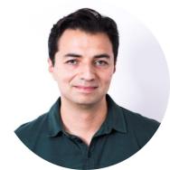 Foto de um homem branco esboçando sorriso. Ele tem olhos claros e cabelos curtos, lisos e escuros. Ele usa camisa polo verde escura e o fundo da imagem é branco. 