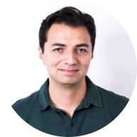 Foto de um homem branco esboçando sorriso. Ele tem olhos claros e cabelos curtos, lisos e escuros. Ele usa camisa polo verde escura e o fundo da imagem é branco. 