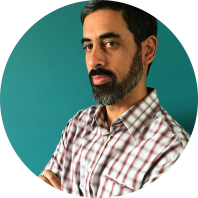 Foto de um homem branco de semi-perfil. Ele tem cabelos curtos e castanhos, olhos castanhos, barba preta e levemente grisalha e veste uma camisa xadrez branca e marrom. O fundo é verde.