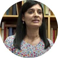 Foto de uma mulher parda falando. Ela tem olhos escuros, cabelos lisos, longos e castanhos. Ela veste uma blusa estampada. No fundo, há uma estante com livros. 