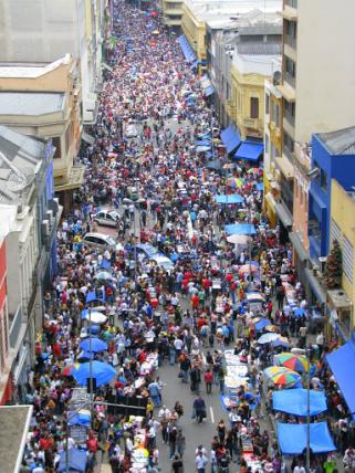 Foto, a partir do alto, de rua cheia de pessoas e barracas de venda, sendo em sua maioria lonas e guarda-sóis azuis. É dia, há alguns carros em meio às pessoas e poucos espaços livres para circulação. Em ambos os lados da rua há edifícios, alguns com três andares e outros mais altos.