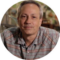 Foto de homem branco esboçando um sorriso. Ele tem olhos castanhos e cabelos curtos, lisos e grisalhos. Usa camisa xadrez cinza e vermelha. O fundo está desfocado. 