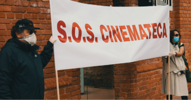 Foto de duas pessoas em frente à Cinemateca. Elas seguram uma faixa branca com a escrita vermelha: "S.O.S. CINEMATECA".