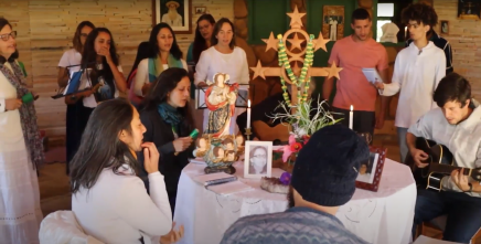 Print de um trecho do documentário Natureza e a comunidade daimista do Céu do Gamarra: conexão com o lugar. No fundo, várias mulheres, todas usando alguma roupa branca, formam um coro. No meio da imagem, há uma mesa com uma toalha branca, sobre a qual está o retrato de uma mulher, duas velas acesas, uma imagem de um santo e um Santo Cruzeiro, símbolo da doutrina. Ao redor da mesa, estão duas mulheres e três homens sentados, dos quais dois tocam violão. Ao fundo, mais três homens encontram-se de pé.
