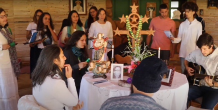 Print de um trecho do documentário Natureza e a comunidade daimista do Céu do Gamarra: conexão com o lugar. No fundo, várias mulheres, todas usando alguma roupa  branca, formam um coro. No meio da imagem, há uma mesa com uma toalha branca, sobre a qual está o retrato de uma mulher, duas velas acesas, uma imagem de um santo e um Santo Cruzeiro, símbolo da doutrina. Ao redor da mesa, estão duas mulheres e três homens sentados, dos quais dois tocam violão. Ao fundo, mais três homens encontram-se de pé.