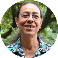 Foto de mulher negra sorrindo. Ela tem olhos castanhos, cabelos longos e escuros, que estão presos. Usa óculos de armação fina arredondada, corrente prata e camisa estampada em tons de azul e preto. Atrás dela há vegetação.