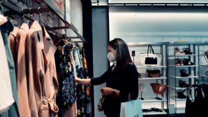 Foto de uma mulher escolhendo roupas em uma loja. A mulher é branca, possui cabelos lisos e escuros e está usando máscara. A roupa dela é preta e ela está com um relógio no braço esquerdo, no qual pendura uma sacola de compras. Essa mulher está escolhendo roupas entre várias opções penduradas em uma arara. Ao fundo, pode-se ver prateleiras com bolsas e sapatos.