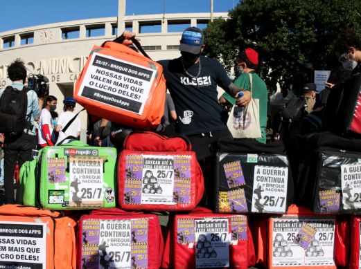 Fotografia da greve dos aplicativos, realizada em 2020. No primeiro plano estão diversas bags (bolsas térmicas dos entregadores), nas cores vermelho, verde, laranja e preto. Cartazes de mobilização da greve estão colados nas bags. Ao centro da imagem, um entregador de camiseta preta e boné preto segura uma bag laranja com um cartaz que diz “Nossas vidas valem mais que o lucro deles!”. Ao fundo se reúnem outras pessoas em frente ao estádio municipal do Pacaembu, em São Paulo.