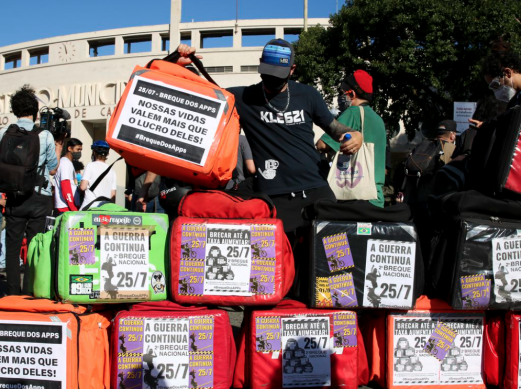 Fotografia da greve dos aplicativos, realizada em 2020. No primeiro plano estão diversas bags (bolsas térmicas dos entregadores), nas cores vermelho, verde, laranja e preto. Cartazes de mobilização da greve estão colados nas bags. Ao centro da imagem, um entregador de camiseta preta e boné preto segura uma bag laranja com um cartaz que diz “Nossas vidas valem mais que o lucro deles!”. Ao fundo se reúnem outras pessoas em frente ao estádio municipal do Pacaembu, em São Paulo. 