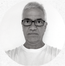 Foto em preto e branco de um homem branco, idoso e de cabelos brancos vestindo uma camiseta branca básica e óculos pretos quadrados.