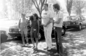 Foto em preto e branco de quatro pessoas em pé, duas mulheres e dois homens. Eles estão na rua e em seu redor há carros estacionados e árvores. Uma das mulheres apoia uma câmera antiga em seu ombro e olha pelo visor com um de seus olhos. A outra mulher segura alguns cabos.