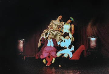 Foto de quatro pessoas apresentando uma peça de teatro. Duas estão sentadas em um sofá vermelho e olham para o lado direito, elas vestem ?. Atrás do sofá, duas personagens usando vestidos longos e coloridos estão de braços dados. Duas cadeiras aparecem mais ao fundo. A iluminação do palco está escura.