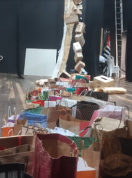 Foto da obra de arte vista de lado. Em primeiro plano, aparecem diversas sacolas de papel agrupadas. Mais ao fundo, está uma cortina de papéis reciclados e uma espécie de corrente de caixas de papelão amarradas umas às outras.