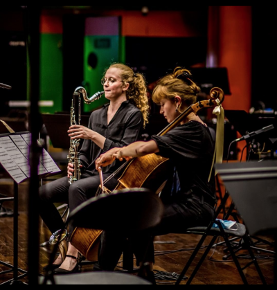 Foto de duas jovens instrumentistas brancas. Uma tem cabelos loiros ondulados, usa óculos e toca saxofone. A outra tem cabelos loiros lisos e presos e toca um violoncelo. Ambas estão vestidas de preto e sentadas diante de um suporte com partitura. Ao redor há cadeiras pretas e suportes de partitura vazios, além de pedestais de microfones. Ao fundo, luzes em tons de vermelho, verde e roxo.