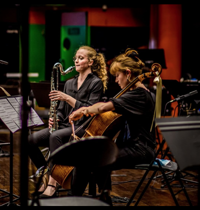 Foto de duas jovens instrumentistas brancas. Uma tem cabelos loiros ondulados, usa óculos e toca saxofone. A outra tem cabelos loiros lisos e presos e toca um violoncelo. Ambas estão vestidas de preto e sentadas diante de um suporte com partitura. Ao redor há cadeiras pretas e suportes de partitura vazios, além de pedestais de microfones. Ao fundo, luzes em tons de vermelho, verde e roxo. 