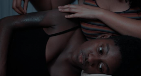 Janaína, moça negra de pele retinta e cabelos curtos e crespos, está deitada e apoia a cabeça no colo de outra moça, com tom de pele mais claro e de quem só vemos o colo e parte dos braços e mãos. Ela acaricia o braço direito de Janaína, que está com um hematoma.