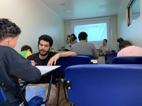 Foto de diversas pessoas sentadas em uma sala de aula. Em primeiro plano, um jovem está agachado diante de uma carteira e conversa com um rapaz que está sentado. Ao fundo, uma jovem manuseia uma câmeras fotográfica e um homem, o professor Marciel Consani, fala segurando um refletor ring light. Os demais jovens, sentados em carteiras, observam e fazem anotações.