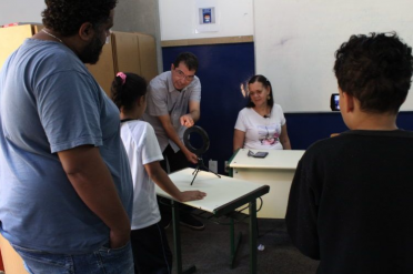 Foto do professor Marciel Consani durante oficina de fotografia. Ele é branco, magro, tem cabelos escuros e usa óculos. Ele explica para uma criança e um jovem negros o uso de um refletor ring light de mesa. Ao fundo, uma mulher branca de cabelos loiros está sentada em uma carteira, posicionada diante de uma câmera fotográfica que é operada por um outro jovem negro participante da oficina.