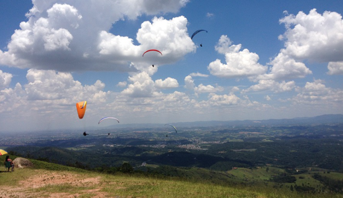 Foto da vista panorâmica de uma região de Mogi das Cruzes. A imagem é formada por morros verdes, céu azul e nuvens. Algumas pessoas voam de parapente.