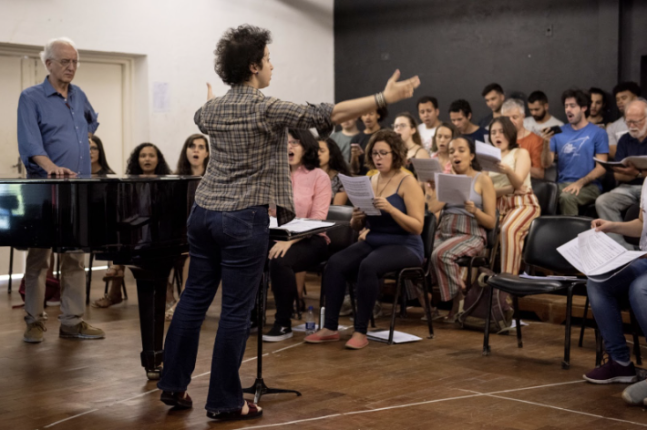 Foto do ensaio do Coral da ECA-USP. À frente do Coral, Laura Gentile de perfil, uma mulher branca, de cabelos curtos escuros, calça jeans e sandálias, fazendo um gesto de regência. Diante dela está um coro de cerca de 25 pessoas sentadas, sendo as duas primeiras fileiras compostas por mulheres e, nas cadeiras mais ao fundo e mais altas, homens. Do lado esquerdo da regente, um piano preto e o professor Marco Antonio da Silva Ramos , que está em pé com a mão direita apoiada no piano. Ele é um homem branco, idoso de cabelos brancos, camisa azul, calça e sapatos marrons.