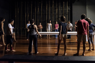 Foto de cerca de 15 pessoas em círculo em um palco de madeira. No meio do círculo há uma mesa de cor branca retangular. O palco está com luz baixa e as paredes ao fundo são pretas e cinza.