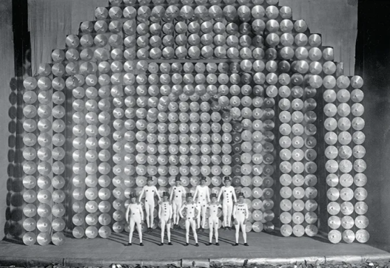 Foto em preto e branco de um palco com um grupo de pessoas vestindo macacões brancos com estampa de bolas e usando chapéus escuros. O palco é decorado com uma estrutura composta por várias fileiras de discos que formam arcos ao fundo, sendo os do centro em maior profundidade. No centro do palco,cinco pessoas estão de pé e outras quatro se elevam do chão apoiando as mãos sobre os ombros dos primeiros.