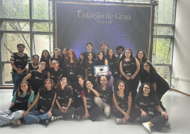 Foto de 23 pessoas, em sua maioria jovens e brancas, dispostas em pé e sentadas, vestidas com coletes pretos e sorrindo para a câmera . Uma garota de cabelos escuros segura uma placa com os dizeres “Missão Cumprida”. Ao fundo há um painel azul escuro onde está escrito em dourado “Colação de Grau ECA-USP” e atrás há uma janela de vidro que vai do chão ao teto.