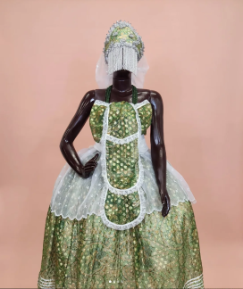 Foto de um manequim escuro vestido com uma indumentária tradicional, composta por um vestido longo verde com detalhes dourados e renda branca. O traje inclui um avental decorativo e um acessório de cabeça adornado com pedrarias e franjas prateadas que cobrem o rosto. O fundo é em tom bege.