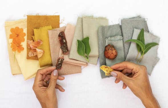 Foto de tecidos de diferentes tons organizados sobre uma superfície branca. Elementos naturais, como flores, folhas, cascas de árvore e frutos, estão dispostos sobre os tecidos. Duas mãos seguram materiais naturais, associando-os às cores dos tecidos.