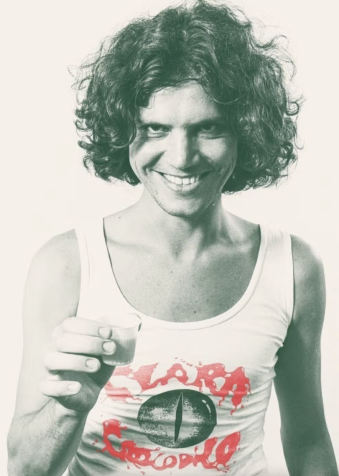 Foto de uma pessoa jovem com cabelo cacheado e volumoso sorrindo diretamente para a câmera enquanto segura um pequeno copo. Veste uma regata branca com a estampa "Clara Crocodilo" em letras vermelhas e o desenho de um olho estilizado no centro. A imagem tem aparência retrô com tonalidade esverdeada.