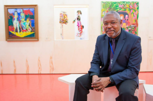 o artista Claudinei da Silva é um homem negro, cabelos curtos grisalhos, com óculos de armação arredondada. Esta sentado em um banco e veste terno azul marinho e gravata. Ao fundo da imagem, observam-se três quadros na parede, obras de arte coloridas e levemente desfocadas.