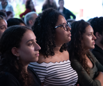 Foto do perfil de três mulheres. As jovens estão sentadas uma ao lado da outra e olham na mesma direção. Ao redor delas, diversas pessoas estão sentadas também. Ao fundo, outras pessoas estão em pé.