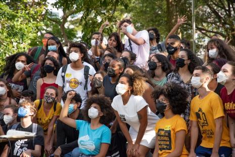 Foto de muitos jovens agrupados, alguns sentados e outros em pé. São meninos e meninas, brancos e negros, que posam para uma outra câmera. A grande maioria deles usa máscara e alguns estão vestindo camisetas de entidades estudantis.