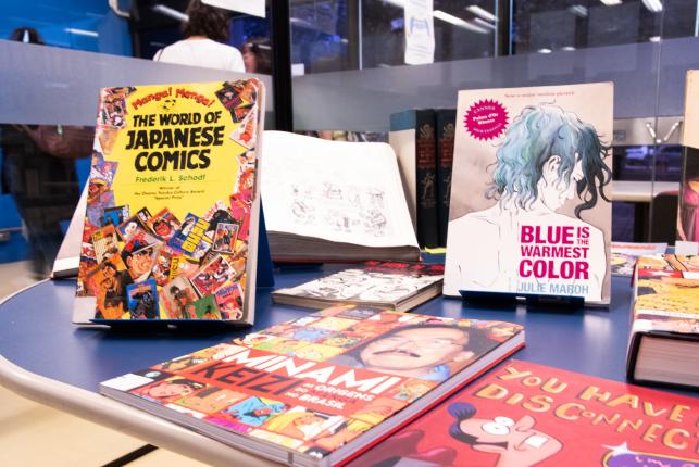 Fotografia de revistas de mangá sobre uma mesa azul, sendo alguns deitados e outros na vertical. Alguns dos títulos são: The World of Japanese Comics, Minami Keizi, Blue is the warmest color, You have disconnection. No fundo, portas de vidro fechadas e pessoas no outro ambiente.