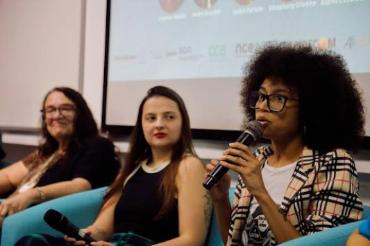 Foto de três mulheres sentadas lado a lado. Da direita para a esquerda: uma mulher negra, de cabelos crespos e castanhos, olhos castanhos, segurando um microfone e vestindo uma camiseta branca estampada, blazer xadrez e óculos de armação preta; uma mulher branca, de cabelos lisos, castanhos e longos, vestindo uma regata preta e usando batom vermelho; uma mulher branca, de cabelos lisos, castanhos e longos, usando um óculos de armação marrom e vestindo uma camiseta estampada e preta. Todas estão sentadas em poltronas azuis. Ao fundo há uma parede branca, com uma projeção. 