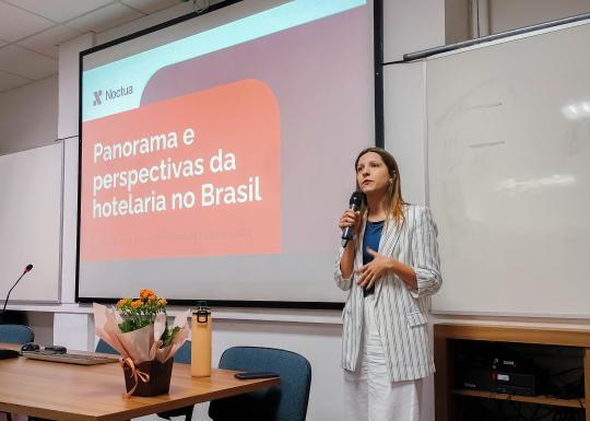 Foto de uma mulher branca em pé falando ao microfone. Ela tem cabelos castanhos claros e usa uma camiseta azul com calça e blazer brancos. No fundo, uma projeção com o título “Panoramas e perspectivas da hotelaria no Brasil”.  À frente da mulher, uma mesa de madeira com um teclado, uma garrafa, um celular e flores. 