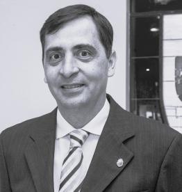 Foto em preto e branco de um homem sorrindo. Ele é branco, tem cabelos lisos e curtos, veste terno, camisa e gravada. Ao fundo, um vitral e uma parede branca.